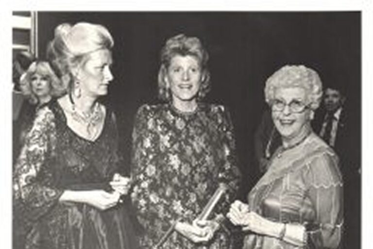 Three women in formal attire stand together, smiling, at what appears to be a formal event or gathering.