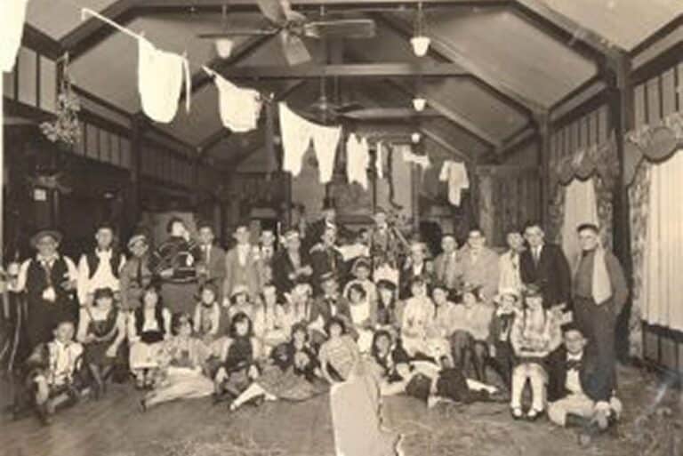 Large group of people in vintage attire pose in a hall with laundry hanging from the ceiling, likely at a themed party.