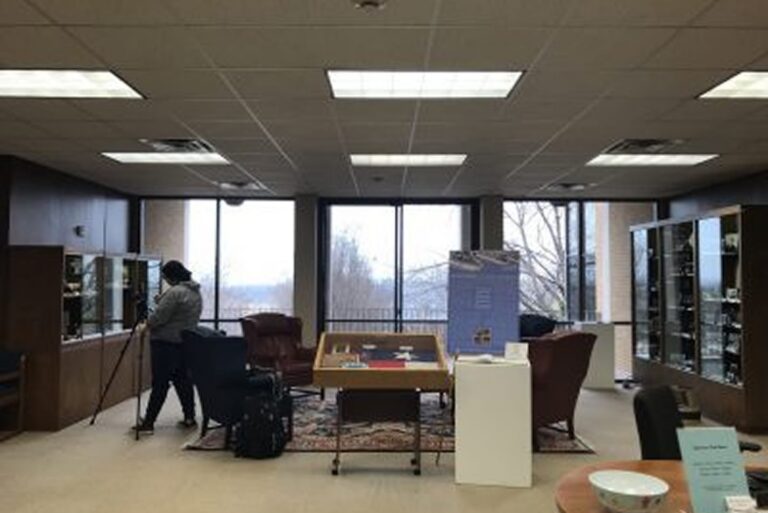 A person uses a tripod in a library room with large windows, display cases, and armchairs.