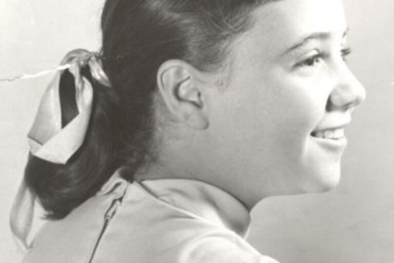 Black-and-white photo of a smiling girl in profile, wearing a ribbon in her hair and a zipper-up top.
