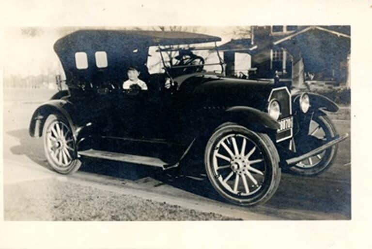 Vintage black car with large spoked wheels parked on street; person sitting in driver’s seat, house in background.