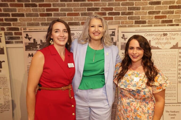 Three women smiling and posing together indoors in front of a brick wall and historical newspaper displays.