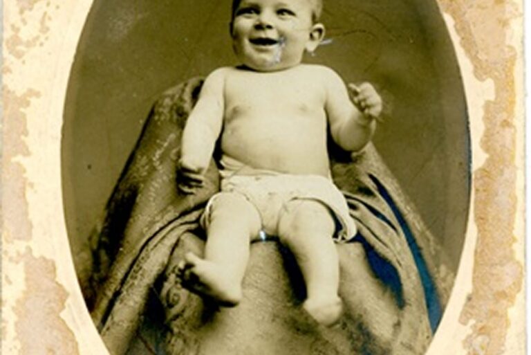 Vintage sepia photo of a smiling baby sitting on a draped lap, partially hidden behind a blanket.