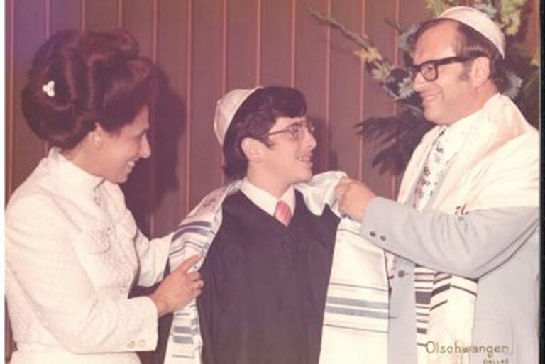A boy in a suit is wrapped in a tallit by two adults, all smiling, during a Jewish ceremony.