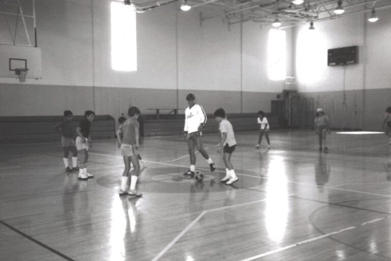 Seven kids and an adult play soccer in a gymnasium with sunlight streaming through tall windows.