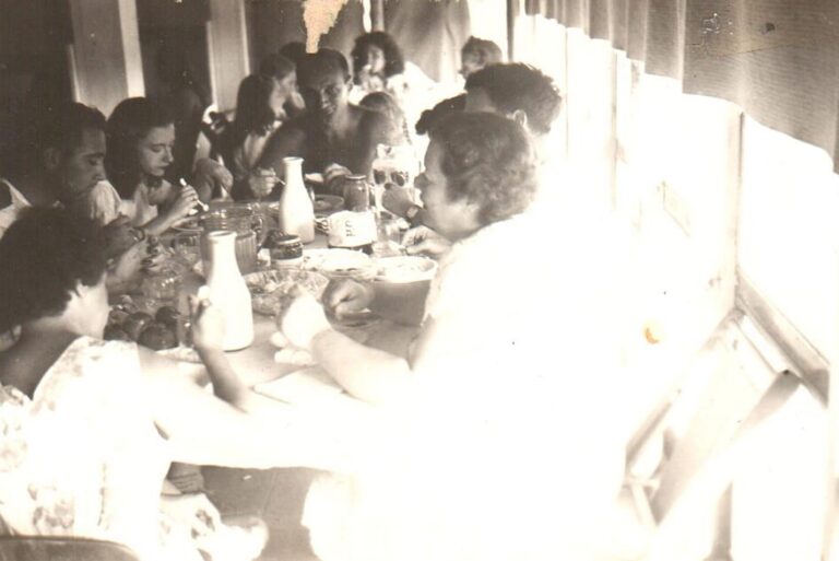 A group of people sit around a table, eating and talking by a bright window in a vintage black-and-white photo.