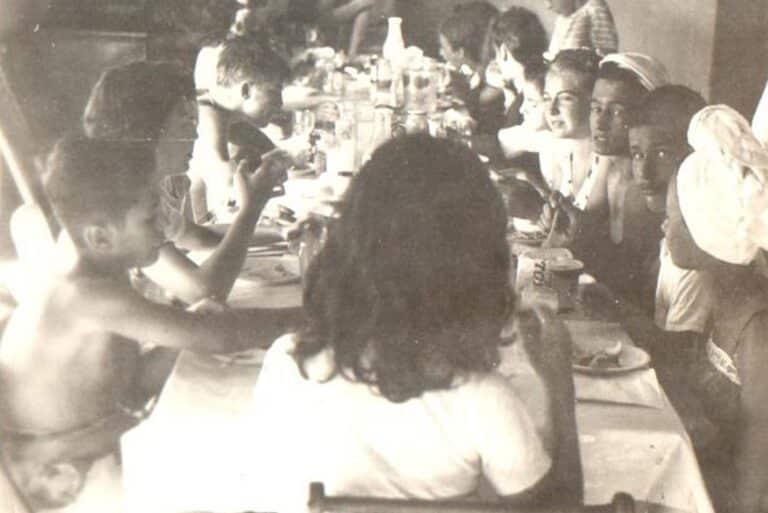 Children and adults sit around a long table, sharing a meal in a warm, lively, black-and-white scene.