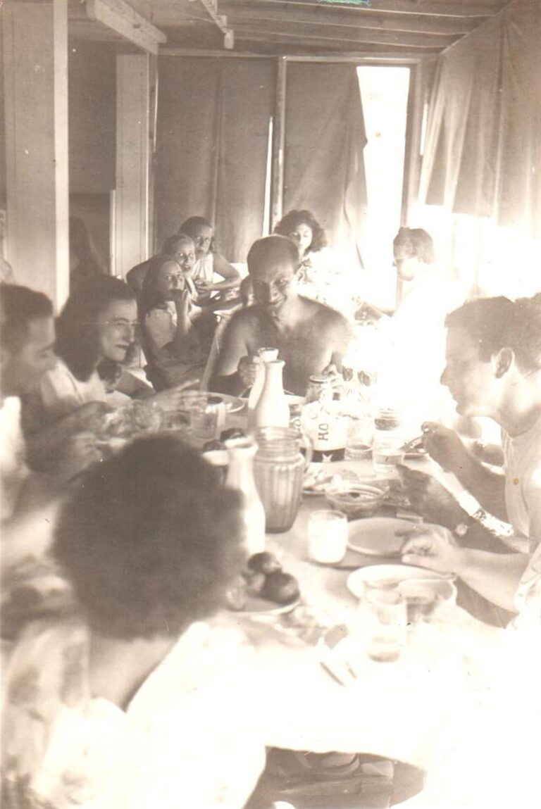 A group of people share a meal around a long table in a sunlit room, smiling and talking together.