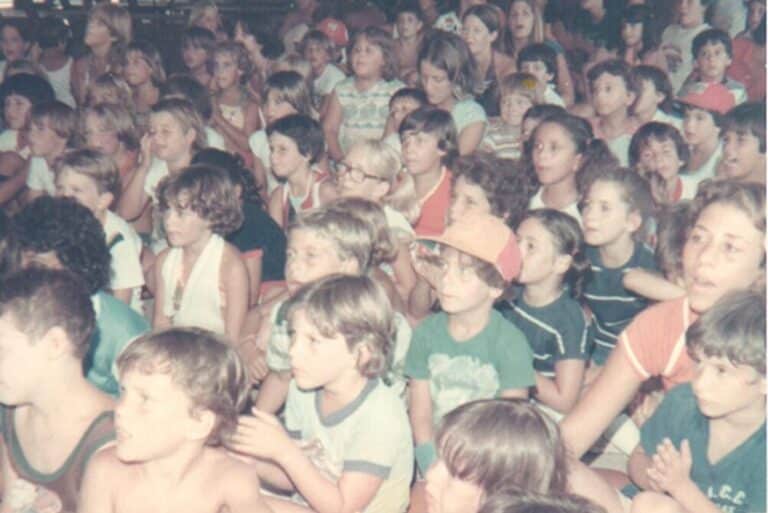 Large group of children sitting closely together, watching something off-camera, wearing summer clothes.