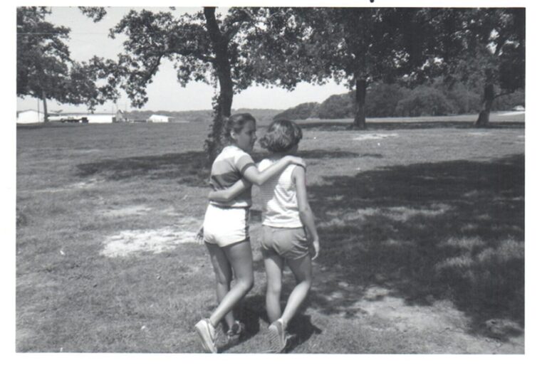 Two people walk arm in arm on a grassy field under trees, seen from behind in a black and white photo.