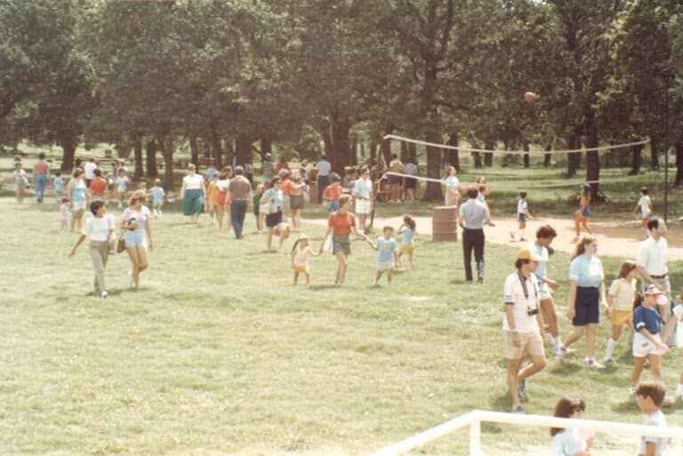 People gather in a sunny park, with children playing and a group near a volleyball net. Trees fill the background.