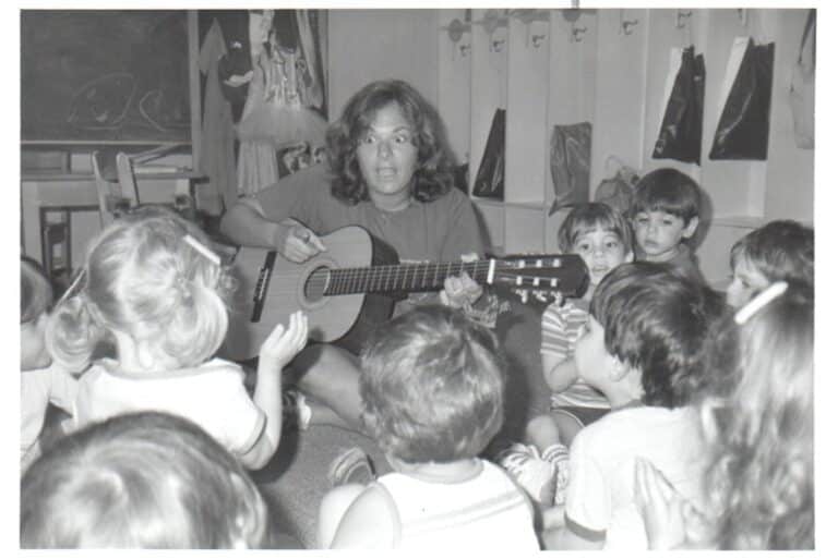 A woman plays guitar and sings to a group of young children seated around her in a classroom.