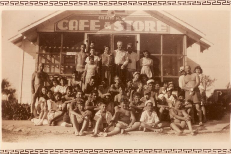 A large group of people pose in front of a vintage cafe and store in a sepia-toned photograph.