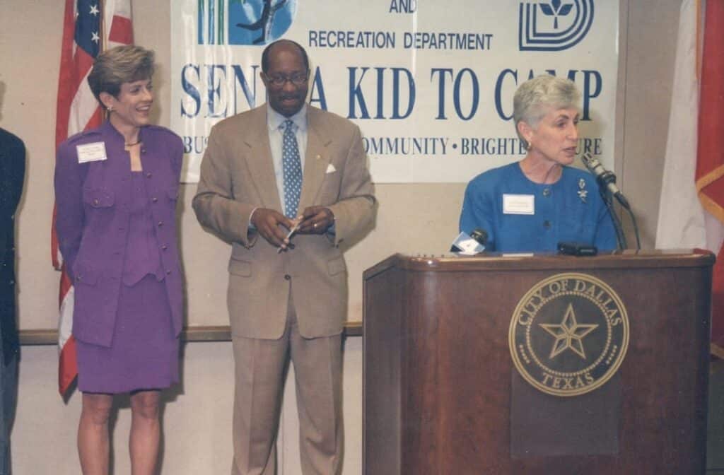 Three people speak at a podium for a “Send a Kid to Camp” event in Dallas, Texas, with flags in the background.