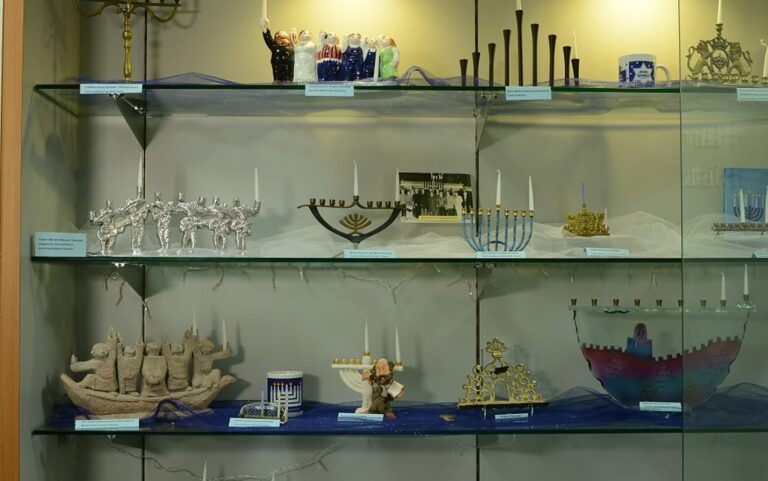 A display case with various menorahs, each uniquely designed, arranged on glass shelves under soft lighting.