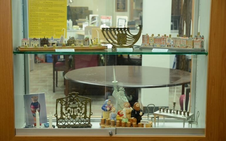 A display case with menorahs, miniature buildings, figurines, and other decorative Jewish items on glass shelves.