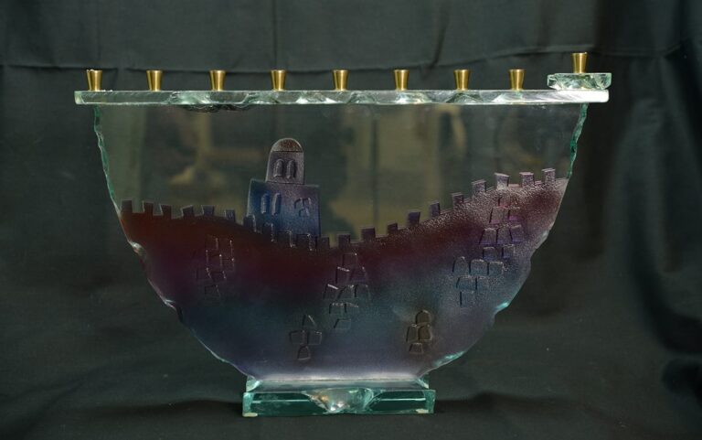 Glass menorah with brass candle holders, featuring a walled city design and a tower, set against a dark background.
