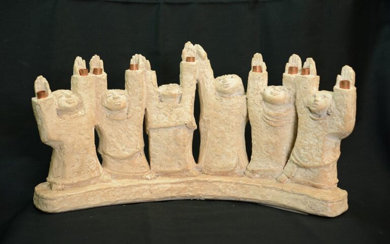 Ceramic sculpture of seven robed figures standing in a row, arms raised, on an arched base, against a black background.