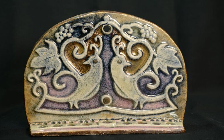 Ceramic art piece with two stylized birds, grape clusters, and leaves in raised relief against a dark background.