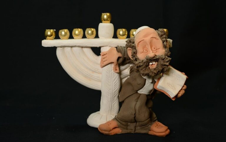 Clay figurine of a smiling man holding a book, standing by a menorah with gold candles, on a black background.