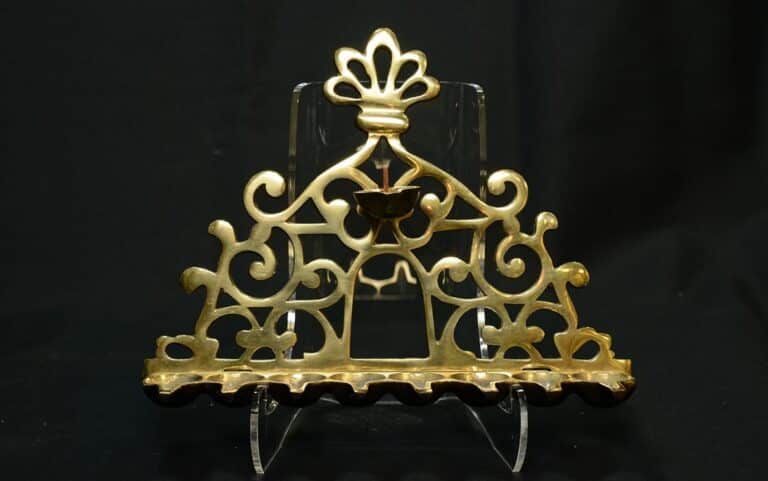 Ornate golden menorah with intricate swirls and a central flower detail, displayed on a clear stand against a black background.