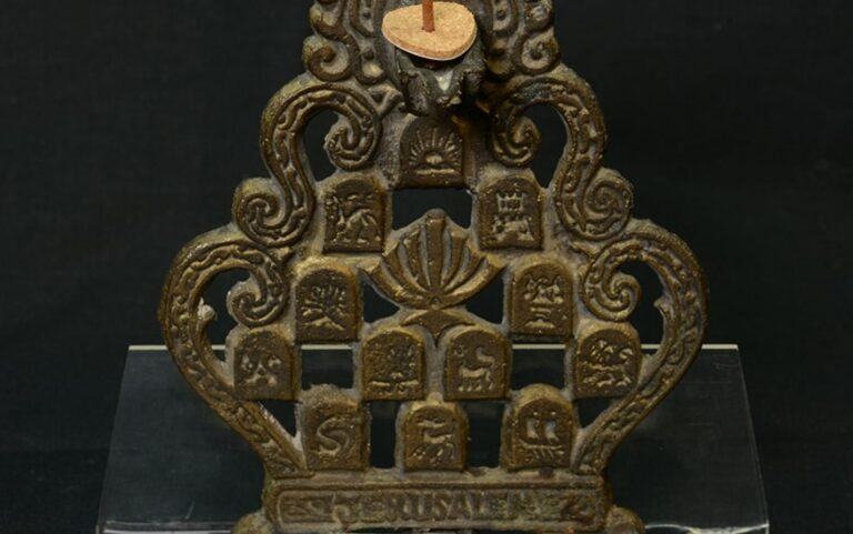 Ornate metal menorah with embossed symbols and candle holder on top, displayed against a black background.