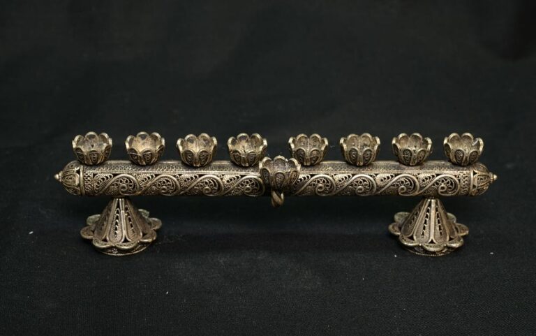 Ornate metal menorah with intricate designs and eight candle holders on a black background.