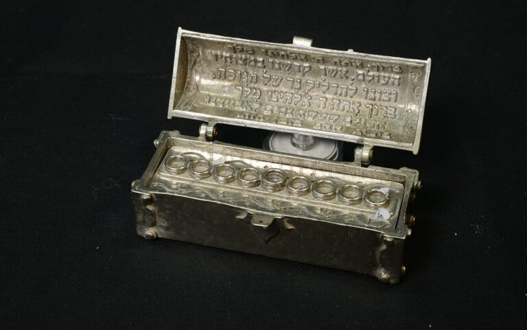 A silver Hanukkah menorah with small glass holders and Hebrew text inside the open lid, set against a black background.