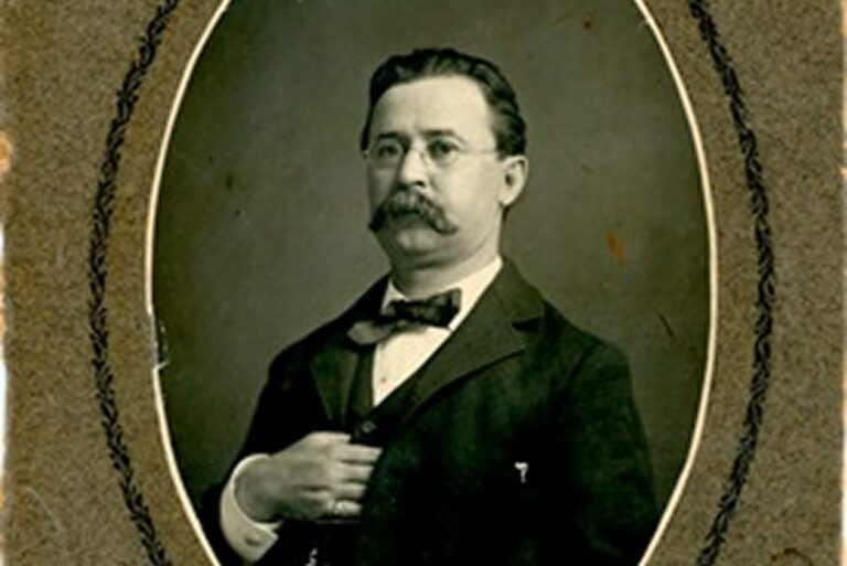 Sepia portrait of a man with glasses, mustache, bow tie, and suit, posing with one hand inside his jacket.