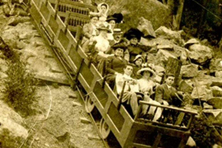 Vintage photo of people in hats and formal clothes riding a wooden incline railway on a rocky hillside.