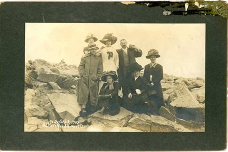 Seven people in early 1900s clothing pose on rocky terrain; some are standing, others are seated.