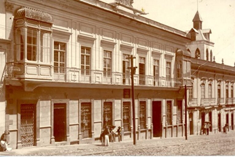 Sepia photo of a historic building with balconies and people standing on a cobblestone street.