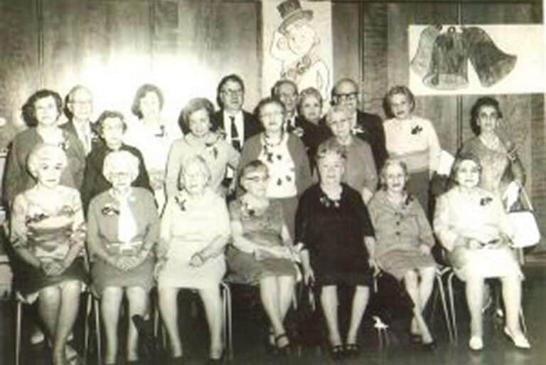 A group of older adults pose for a formal photo indoors, with wall decorations behind them.