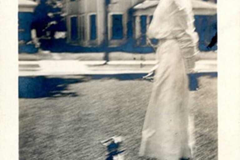 A woman in a long dress stands outside near a house, with a small blurry animal at her feet.