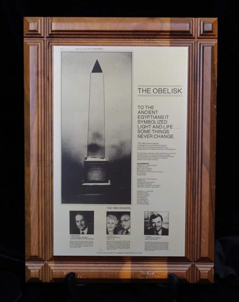 Framed plaque with a photo of an obelisk, text about its symbolism, and small portraits of three individuals at the bottom.
