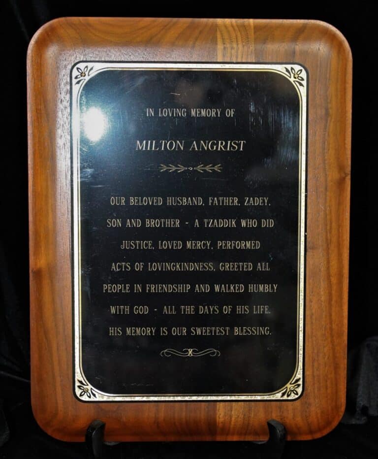 Wooden plaque with an inscription honoring Milton Angrist, describing his kindness and legacy, on a black background.