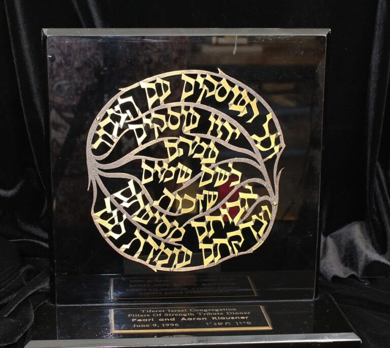 A decorative plaque with intricate Hebrew lettering and a gold and black design, displayed on a black stand.