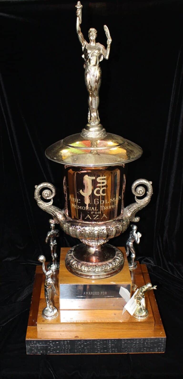Large silver trophy with human figures, two handles, and wooden base, awarded for a memorial tennis event.