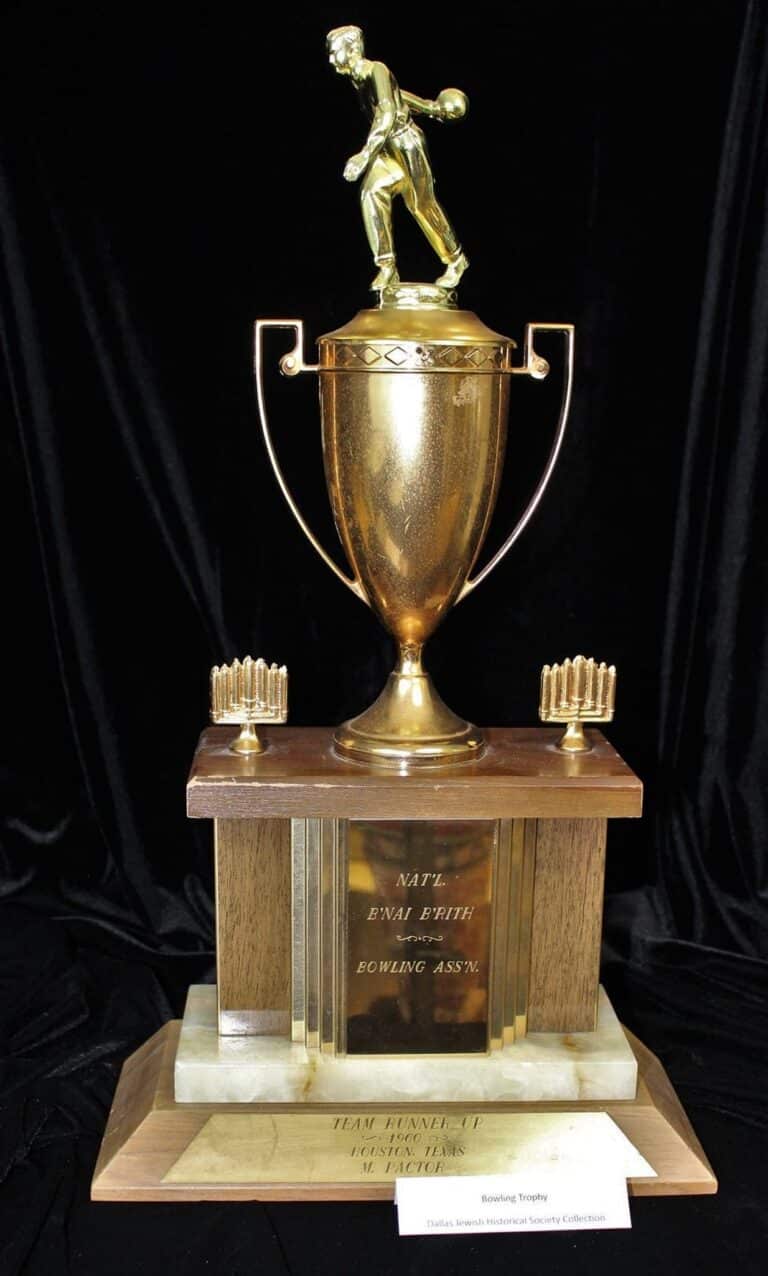 Gold bowling trophy with bowler figure, wooden base, and small candle holders, displayed against a black background.