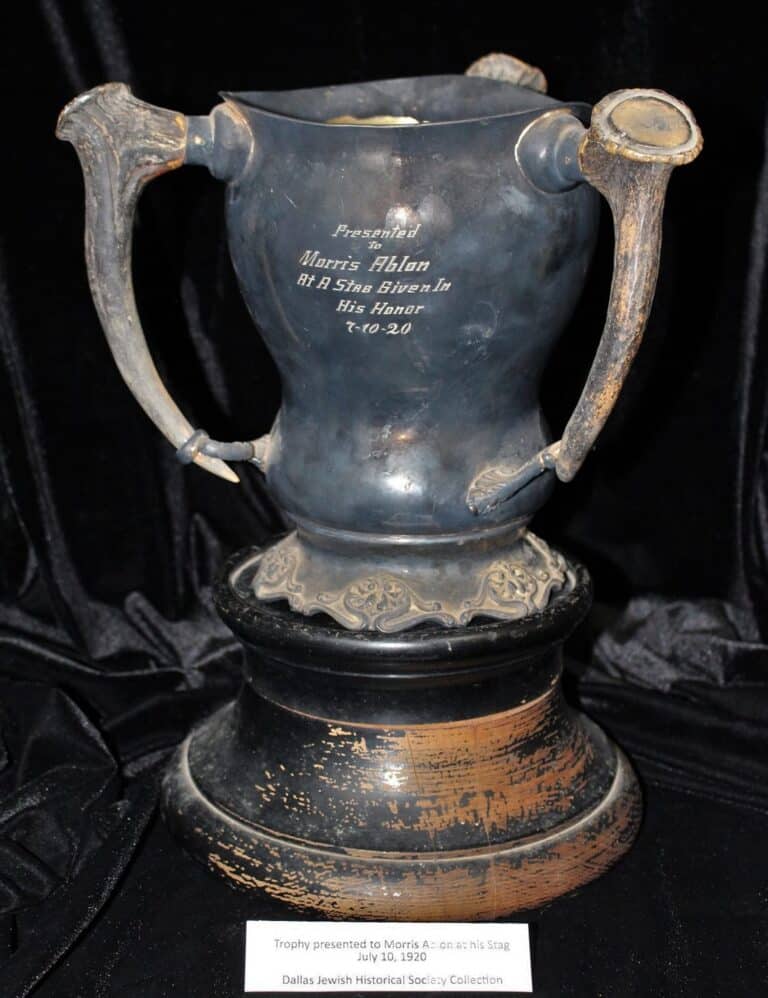 A tarnished trophy cup with ornate handles, engraved plaque, and worn base, displayed on a dark cloth background.