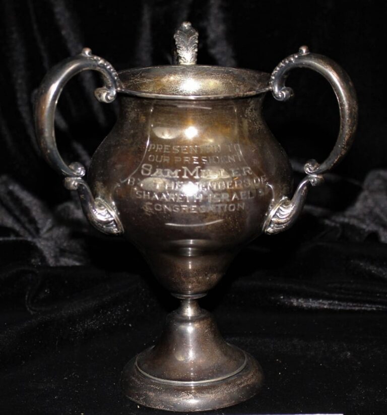 A tarnished silver trophy cup with two handles and engraved text, placed against a dark, velvety background.