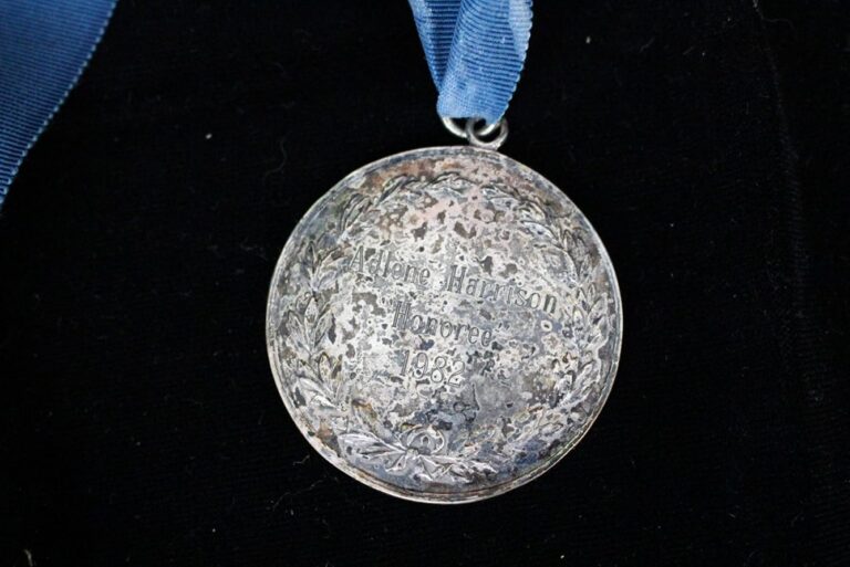 A tarnished silver medal with a blue ribbon, engraved with “Irene Harison, Harrow, 1902” inside a laurel wreath.