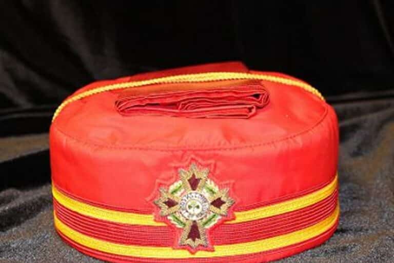 A red ceremonial hat with yellow stripes and an embroidered cross emblem at the front, placed on a dark surface.