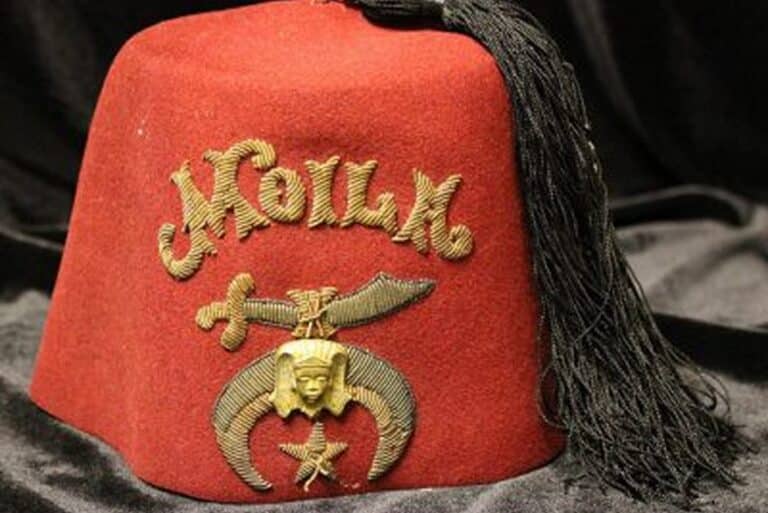 A red fez hat with gold embroidery, a sword, crescent, star, and a tassel on a black cloth background.