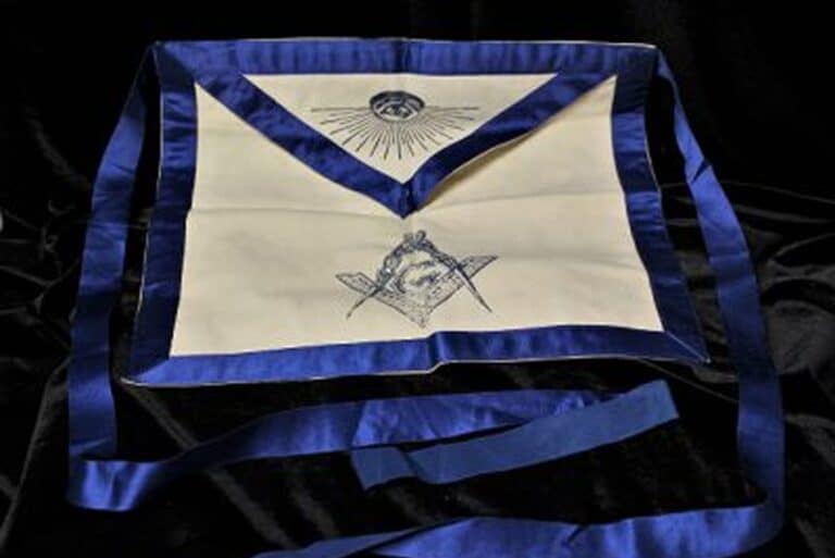 A white Masonic apron with blue trim, featuring Masonic symbols, displayed on a dark fabric background.