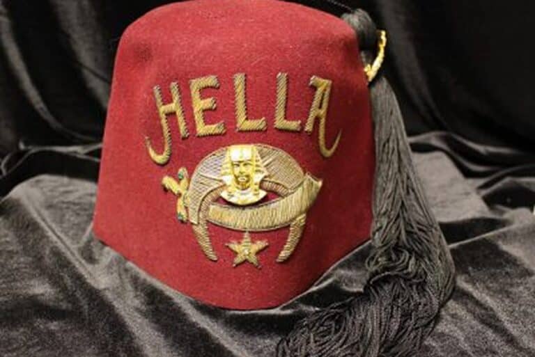 A maroon fez hat with “HELLA” and gold embroidered symbols, including a star, scimitar, and face, plus a black tassel.