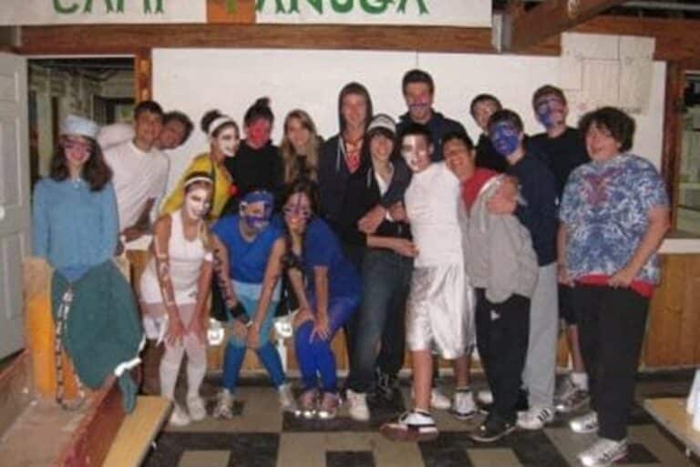 A group of teens with painted faces pose together indoors at what appears to be a camp.