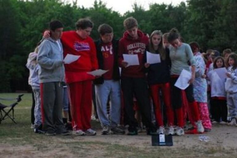 A group of teens stands outdoors in a circle, reading papers, with trees in the background.