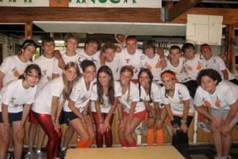 A group of teens in matching white shirts and red shorts pose together and smile inside a rustic building.