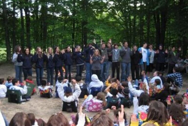 A group of people stands in a line outdoors, facing an audience sitting on the ground in a wooded area.